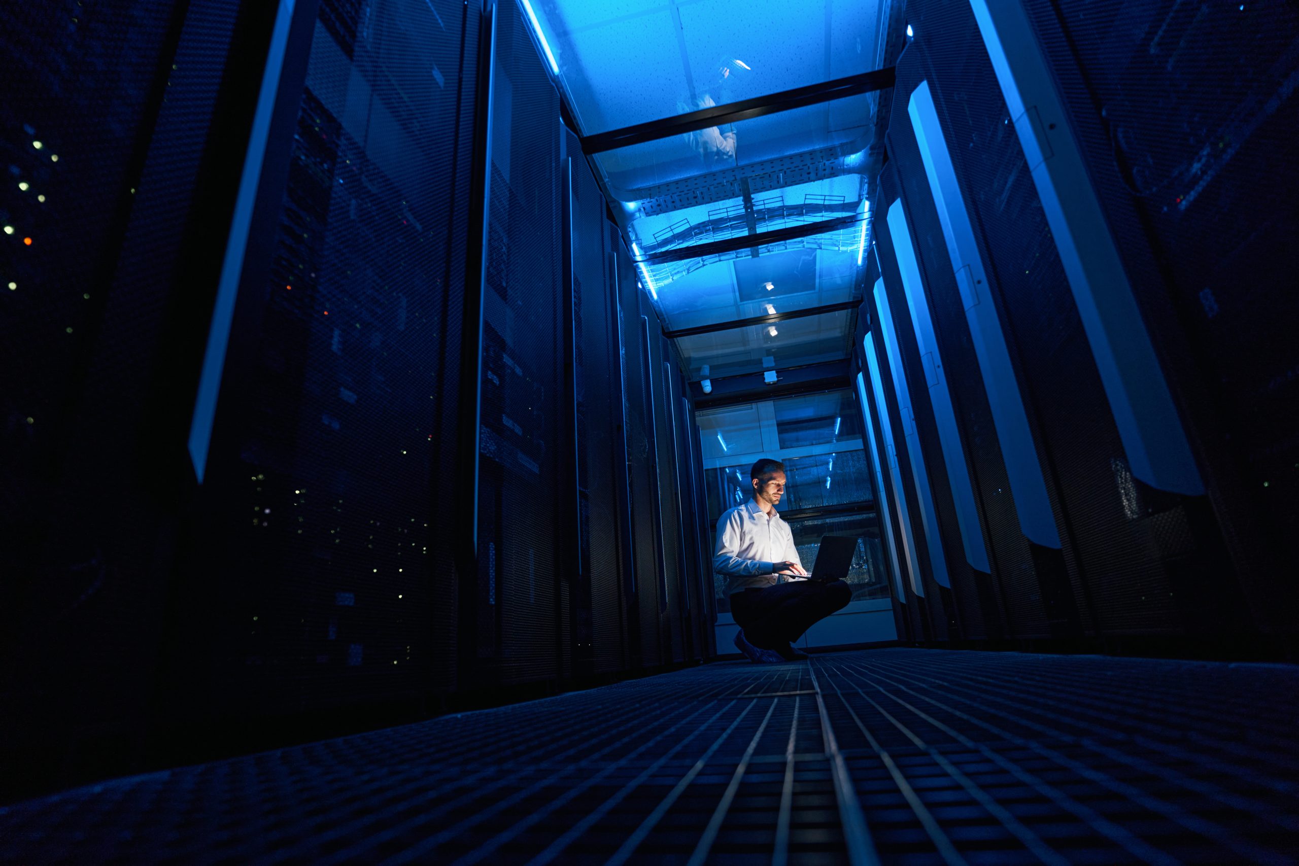 IT specialist sitting in passage between server cabinets and checking data in his laptop